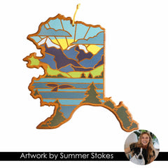 Beautifully crafted from wood, this Alaska-shaped cutting board features a vibrant landscape depicting mountains, trees, and a sunset. It includes a charming circle illustrating a smiling woman with her dog. At the bottom, the text reads "Summer Stokes Artwork." Created by Totally Bamboo, this piece is reminiscent of state-shaped cutting boards in its precision and charm.