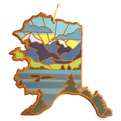 A cutting board from Totally Bamboo, shaped like the state of Alaska, showcases a vibrant landscape with mountains, a lake, pine trees, a yellow sun, and blue skies. The design features cut-out sections that evoke a stained-glass look similar to Summer Stokes' artwork. It includes a looped string at the top for hanging.