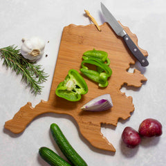 Sliced green bell peppers and a piece of red onion rest on the Alaska Cutting Board with Artwork by Summer Stokes, beautifully crafted from bamboo by Totally Bamboo. Nearby, whole garlic, rosemary, two red potatoes, and cucumbers enhance the scene. A knife sits on this functional and stylish surface, adding both utility and artistic flair to your kitchen.