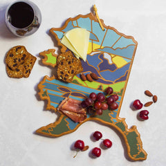 The Alaska Cutting Board with Artwork by Summer Stokes from Totally Bamboo features a vibrant mountain landscape design that artfully presents cheese, cured meat, grapes, and nuts. Accompanying the board are cherries, crackers, and almonds, with a glass of red wine close by on the white surface.