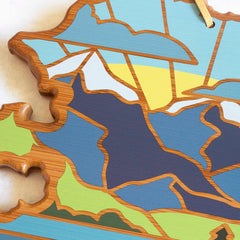 The Alaska Cutting Board by Totally Bamboo features artwork by Summer Stokes, depicting a colorful abstract landscape with a sun-drenched mountain, swirling clouds, and vibrant fields. Its layered illustration bursts with shades of blue, green, yellow, and brown.