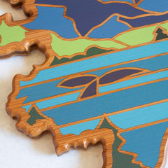 Close-up of an Alaska Cutting Board by Totally Bamboo, decorated with Summer Stokes artwork featuring intricate patterns and abstract, colorful stripes and shapes in blue, green, and brown tones. The design is reminiscent of artistic flair with its wavy edges.