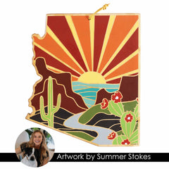 The Arizona Cutting Board from Totally Bamboo is crafted in the shape of the state and showcases a vibrant desert scene with cacti, mountains, and a river beneath a vivid sunrise. A small circular photo depicts someone smiling with a dog. Text on the board reads "Artwork by Summer Stokes.