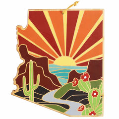 The Arizona Cutting Board with Artwork by Summer Stokes, produced by Totally Bamboo, showcases a stylized illustration of the state of Arizona. The design features a desert landscape on a bamboo cutting surface, complete with elements such as a sunset, cacti, mountains, and a winding river. This state-shaped cutting board is brought to life with bold colors like orange, yellow, and green.