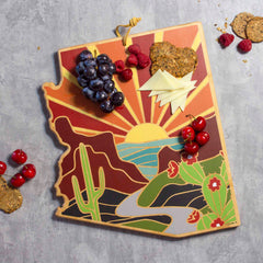 The Arizona Cutting Board with Artwork by Summer Stokes from Totally Bamboo features a vibrant depiction of a desert scene, including cactus and sunset elements. This state-shaped board beautifully holds an assortment of grapes, cheese slices, cherries, and crackers on its bamboo surface. It's ideal for enthusiasts of state-inspired cutting boards and is set against a textured gray background.