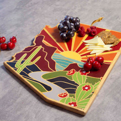 The Arizona Cutting Board with Artwork by Summer Stokes from Totally Bamboo features a vibrant bamboo cutting surface with a desert landscape design, showcasing a sun setting behind mountains. It's beautifully adorned with grapes, raspberries, cherries, cheese slices, and bread on a gray surface.