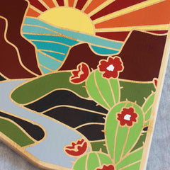 The Arizona Cutting Board with Artwork by Summer Stokes from Totally Bamboo beautifully portrays a sunset over mountains with a flowing river. Blooming red flowers and green cactus in the foreground enhance the scene's charm, while warm yellows, reds, and greens infuse the piece with vibrant energy.