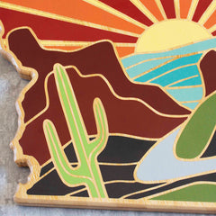 The Arizona Cutting Board by Totally Bamboo showcases a vibrant mural by Summer Stokes, featuring a stylized desert scene with a saguaro cactus in the foreground. The backdrop highlights mountains, a river, and a setting sun with bold colors such as brown, green, blue, and orange radiating across the artwork.