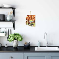 A modern kitchen showcases a black countertop, gray cabinets, and a silver faucet. The counter is adorned with the Arizona Cutting Board from Totally Bamboo, featuring desert-themed artwork by Summer Stokes with sunlit mountains and cacti, along with bamboo utensils. This vibrant artwork also graces the white wall as decorative elements. Shelves display a teapot and cup.