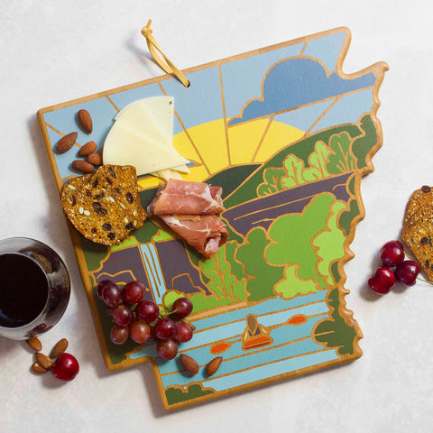 The Arkansas Cutting Board with Artwork by Summer Stokes, from Totally Bamboo, holds cheese, prosciutto, almonds, and crackers. Nearby sit grapes and a glass of red wine, all elegantly displayed on a light marble surface.