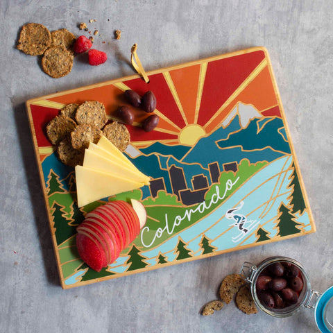 The Totally Bamboo Colorado Cutting Board, adorned with Summer Stokes' artwork, elegantly displays sliced apples, cheese, olives, and crackers on its surface depicting a vibrant Colorado landscape. Beside the board sits a small jar of olives, offering an exquisite touch of nature to your kitchen.