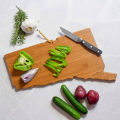 A Connecticut-shaped bamboo cutting board from Totally Bamboo, featuring artwork by Summer Stokes, holds sliced green bell pepper and a knife. Nearby, whole garlic cloves, rosemary sprigs, a cut shallot, two red potatoes, and cucumbers rest on the light-colored background.