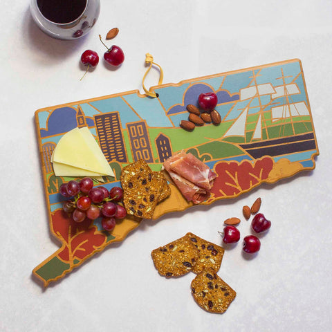 The Connecticut Cutting Board with Artwork by Summer Stokes from Totally Bamboo is a vibrant bamboo surface shaped like the state of Connecticut. It beautifully displays cheese, prosciutto, grapes, and crackers. Scattered almonds and cherries add to the presentation, while a cup of coffee complements the scene from the corner. The board features Summer Stokes' artwork depicting buildings and a ship.