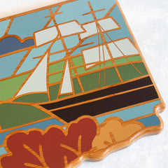 This Connecticut-shaped cutting board by Totally Bamboo, featuring an artwork by Summer Stokes, depicts a stylized ship with sails and incorporates geometric shapes in lively shades of blue, green, brown, and white. Its abstract design gives it a stained-glass window-like appearance.