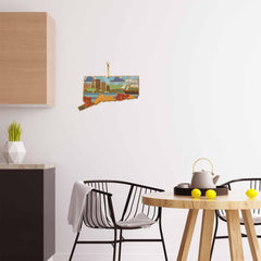 A modern kitchen interior features a round wooden table, two black chairs, and a white teapot resting on the table. The white wall is adorned with a vibrant piece from Summer Stokes Artwork. On the countertop in the background, a green plant is placed beside Connecticut state-shaped cutting boards by Totally Bamboo for an extra touch of charm.