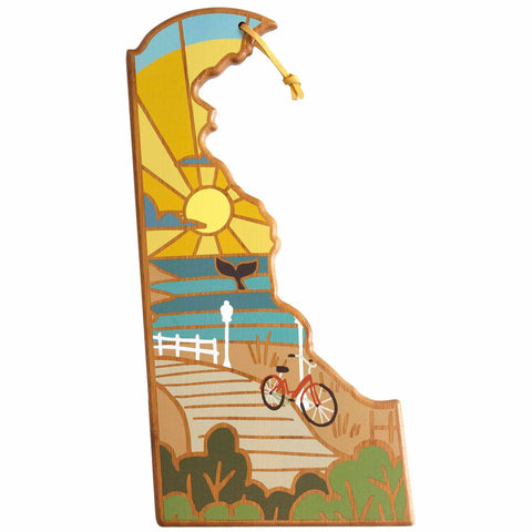 The Delaware Cutting Board with Artwork by Summer Stokes from Totally Bamboo showcases a vibrant illustration of the state of Delaware, highlighting a sunny sky, a beach, a bicycle on a path, and a white fence. The lively design is rich with greenery and blue waves and is elegantly accented with a charming yellow tassel.