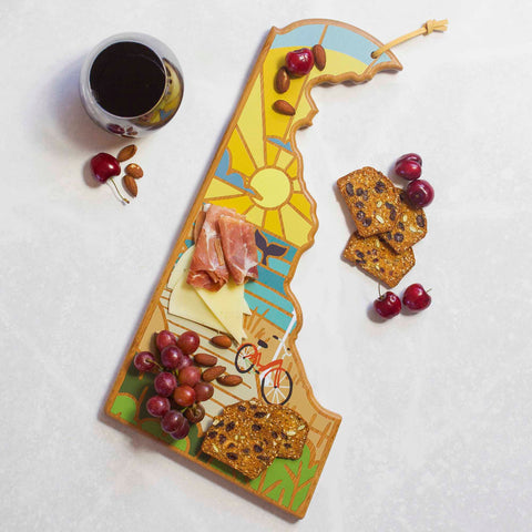 The Delaware Cutting Board with Artwork by Summer Stokes from Totally Bamboo, featuring a sun design, elegantly displays almonds, cherries, crispbreads, cheese, ham, and grapes on its bamboo surface. Nearby on the light-colored surface are scattered cherries and a glass of red wine.