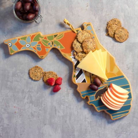 A serving board shaped like Florida, made from bamboo and featuring intricate floral designs, presents an array of cheese slices, apples, olives, and crackers. Raspberries are placed beside it on a gray surface. In the top-left corner sits a jar of olives that enhances the charm of the Totally Bamboo Florida Cutting Board with Artwork by Summer Stokes.