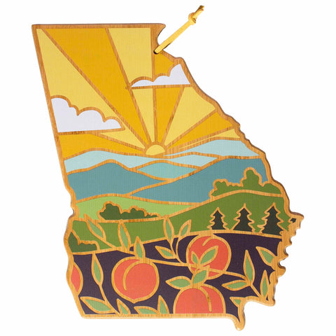 The Georgia Cutting Board with Artwork by Summer Stokes, made by Totally Bamboo, showcases a decorative illustration featuring a sunset over mountains and peach orchards. This state-shaped cutting board has warm colors and stylized landscape elements on its bamboo surface, reminiscent of Summer Stokes' style.