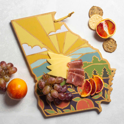 A Georgia Cutting Board with a bamboo surface, displaying cheese slices, prosciutto, grapes, and blood orange slices in an artistic arrangement. Encircled by crackers and an orange, this Totally Bamboo board features Summer Stokes artwork with a vibrant landscape design of sun and mountains.