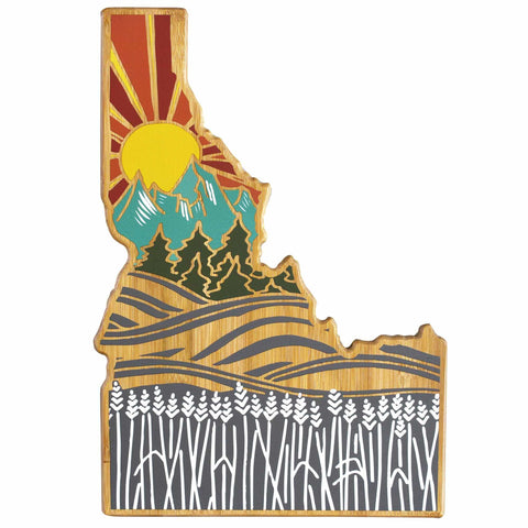 The Idaho Cutting Board with Artwork by Summer Stokes, crafted by Totally Bamboo, beautifully showcases the state's outline. It features a vibrant sunset behind mountains and trees at the top, while the bottom depicts gray and white rolling fields of wheat, highlighting Idaho's agricultural heritage.