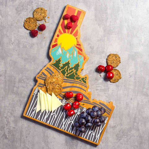 The Idaho Cutting Board by Totally Bamboo, featuring Summer Stokes' vibrant artwork of a sun, mountains, and trees, elegantly displays cheese slices, raspberries, grapes, cherries, and crackers on a gray background.