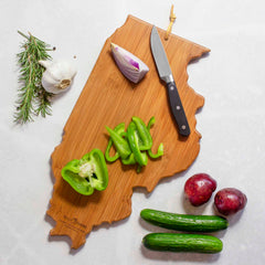 The Illinois Cutting Board with Artwork by Summer Stokes from Totally Bamboo features a bamboo surface styled in the shape of Illinois, showcasing sliced green bell pepper alongside a knife. Complementing this scene are a halved onion, a garlic bulb, rosemary sprigs, two red potatoes, and cucumbers arranged on a light gray surface. An ideal choice for those who cherish state-shaped cutting boards.