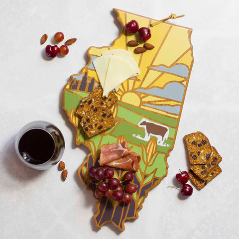 The Illinois Cutting Board with Artwork by Summer Stokes by Totally Bamboo is ideal for displaying a selection of snacks, including cheese slices, prosciutto, grapes, cherries, almonds, and crackers alongside a bowl of dark liquid. Its charm is elevated by a farm scene featuring a cow silhouette.