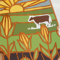 A depiction of a farm scene featuring the sun rising elegantly over expansive fields, inspired by Summer Stokes Artwork. In the distance, a white and brown cow is set against lush green surroundings. Foregrounded corn stalks with mature ears create depth, mirroring the artistic style found on Totally Bamboo’s Illinois Cutting Board with Artwork by Summer Stokes.