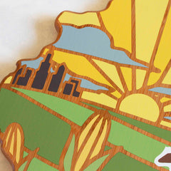 Totally Bamboo's Illinois Cutting Board, featuring artwork by Summer Stokes, showcases a stylized wooden design with a sunburst over a city skyline, accompanied by clouds and fields in the foreground. The vibrant colors of yellow, green, and blue capture the essence of nature at its most lively during summer.