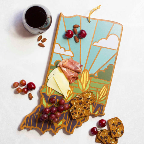 The Indiana Cutting Board with Artwork by Summer Stokes from Totally Bamboo is a decorative piece shaped like the state of Indiana, holding cheese, ham, grapes, and crackers. Almonds and cherries are scattered nearby, accompanied by a cup of coffee at the top left corner. The cutting board features a vibrant landscape illustration created using UV printing techniques.