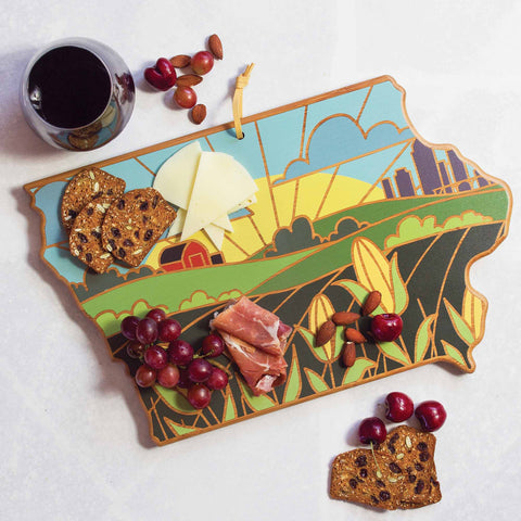 The Iowa Cutting Board by Totally Bamboo, featuring artistic landscape design inspired by Summer Stokes' artwork, is perfect for holding cheese, prosciutto, grapes, almonds, and crackers with a glass of red wine on a light surface.