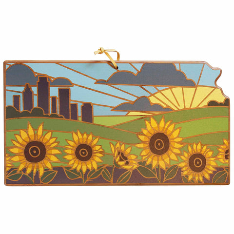 The Kansas Cutting Board, a product by Totally Bamboo, features artwork by Summer Stokes that presents a stylized Kansas landscape with a city skyline, sunrise, and sunflowers. This piece has a stained glass appearance similar to the detailed designs on state-shaped cutting boards and includes a ribbon for hanging.