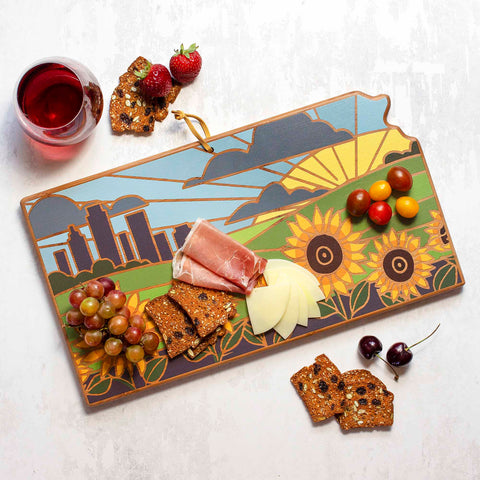 The Kansas Cutting Board with Artwork by Summer Stokes from Totally Bamboo features a beautifully crafted bamboo cutting surface adorned with a vibrant city skyline and sunflower design. It tastefully displays slices of meat and cheese, cherry tomatoes, and grapes next to crackers. Completing the inviting table setting are a glass of red wine, strawberries, and cherries.