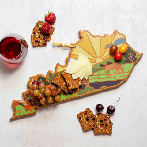 Using the Kentucky Cutting Board with Artwork by Summer Stokes from Totally Bamboo, cheese, grapes, strawberries, and crackers are beautifully arranged. The board showcases a painted design featuring a sunrise, horse, and landscape. Accompanying this arrangement on the light bamboo surface is a glass of red wine with additional crackers nearby.