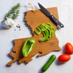 On a Louisiana-shaped cutting board by Totally Bamboo, crafted from a bamboo surface, lies sliced green pepper with a knife. Nearby rest a garlic bulb, rosemary sprig, peppercorns, tomatoes, and a cucumber on a light surface—creating an idyllic scene that brings to mind Summer Stokes' artwork.