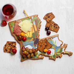 The Louisiana Cutting Board with Artwork by Summer Stokes from Totally Bamboo beautifully showcases cheese, prosciutto, grapes, tomatoes, cherries, and crackers on its surface. Nearby, a glass of red wine and strawberries rest on the textured surface.