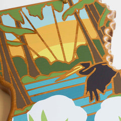 The Louisiana Cutting Board with Artwork by Summer Stokes from Totally Bamboo beautifully captures a stylized landscape at sunrise. The artwork features a blue river winding through the forest, lined with trees, while a heron soars above and fluffy white clouds fill the sky, evoking the serenity of a peaceful morning.