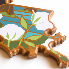 The Louisiana Cutting Board by Totally Bamboo, featuring artwork by Summer Stokes, is a wooden piece shaped like the state. It showcases an illustration of large white flowers with green leaves set against a blue-striped background. The intricately cut edges beautifully outline the state's shape.