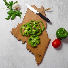 The Totally Bamboo Maine Cutting Board, featuring artwork by Summer Stokes, holds sliced green bell peppers. Nearby, a knife is surrounded by a fresh bell pepper, ripe red tomato, garlic, and sprigs of basil on a light gray surface—bringing to life the essence of Summer Stokes's design.