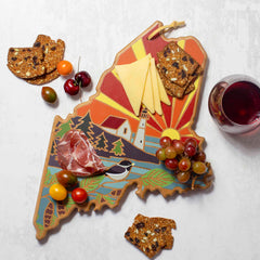 The Maine Cutting Board with Artwork by Summer Stokes from Totally Bamboo displays assorted cheeses, cured meats, and grapes on its colorful, state map-shaped bamboo surface. Nearby are crackers, tomatoes, and cherries alongside a glass of red wine resting on the white surface.