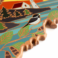 This exquisite Maine Cutting Board by Totally Bamboo features a Summer Stokes artwork, presenting a beautifully carved wooden bird in the foreground, set amidst a vibrant backdrop of forest, river, and house. The design comes alive with bold colors and intricate patterns on the jagged bamboo surface.