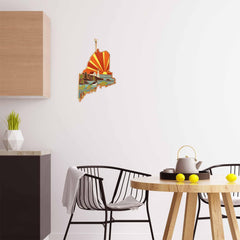 A modern kitchen is adorned with a black chair, a round wooden table, and a white teapot embellished with citrus fruits. On the wall hangs the Maine Cutting Board by Totally Bamboo, featuring Summer Stokes' artwork that captures coastal and sunset hues. The minimalist décor highlights wooden and black elements for a sleek aesthetic.