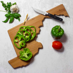 A Maryland-shaped cutting board by Totally Bamboo, featuring artwork by Summer Stokes, cradles sliced green bell peppers. Surrounding it elegantly on a light-colored background are a whole bell pepper, a tomato, garlic clove, basil leaves, and a knife. This composition artfully captures the essence of state-shaped cutting boards.