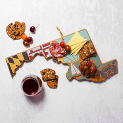 A Totally Bamboo Maryland Cutting Board with Artwork by Summer Stokes elegantly displays a variety of foods such as sliced meats, cheese, grapes, multigrain crackers, and cherry tomatoes. Next to it, a glass of red wine sits on a textured surface.