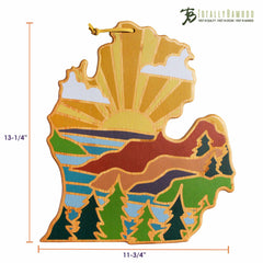 Introducing the Michigan Mitten Cutting Board by Totally Bamboo, featuring Summer Stokes' artwork. This bamboo cutting surface, shaped like Michigan's lower peninsula, displays a stunning sunburst along with water, trees, and clouds. Measuring 13-1/4 inches tall and 11-3/4 inches wide, it's a distinct addition to any collection of state-shaped cutting boards.