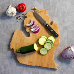 The Michigan Mitten Cutting Board with Artwork by Summer Stokes from Totally Bamboo displays a knife, sliced zucchini, red onion, whole tomato, and garlic cloves on a gray surface. It's an ideal way to showcase your love for state-shaped cutting boards while adding flair to your meal prep.