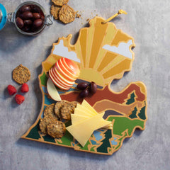 A vibrant, nature-inspired cutting board from Totally Bamboo, shaped like Michigan’s Upper Peninsula and featuring artwork by Summer Stokes, holds sliced cheese, apple pieces, and olives. Crafted with a bamboo cutting surface, it's ideal for snacks. Nearby on a gray surface are crackers and raspberries.