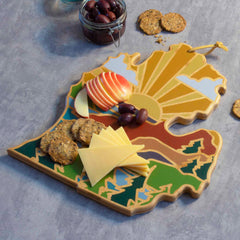 The Michigan Mitten Cutting Board with Artwork by Summer Stokes, from Totally Bamboo, showcases sliced apples, cheese, olives, and crackers on its bamboo cutting surface. The board features vibrant geographic designs reminiscent of State-Shaped Cutting Boards, with a jar containing extra olives nearby on a gray surface.