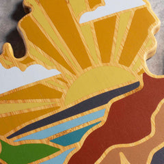 The Michigan Mitten Cutting Board by Totally Bamboo features an artistic design by Summer Stokes that evokes tranquil landscapes. The board beautifully showcases a sunrise with yellow and orange sunrays lighting up clouds, a blue river, green fields, and brown mountains, bringing vibrant color and texture to the piece.